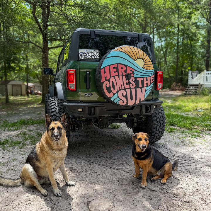 An Eruption Green Ford Bronco is shown from the rear in a wooded outdoor setting, with two dogs sitting on the ground in front of it. The Ford Bronco has a large circular spare tire cover on the back with a retro-style sun and wave design and the text “HERE COMES THE SUN.